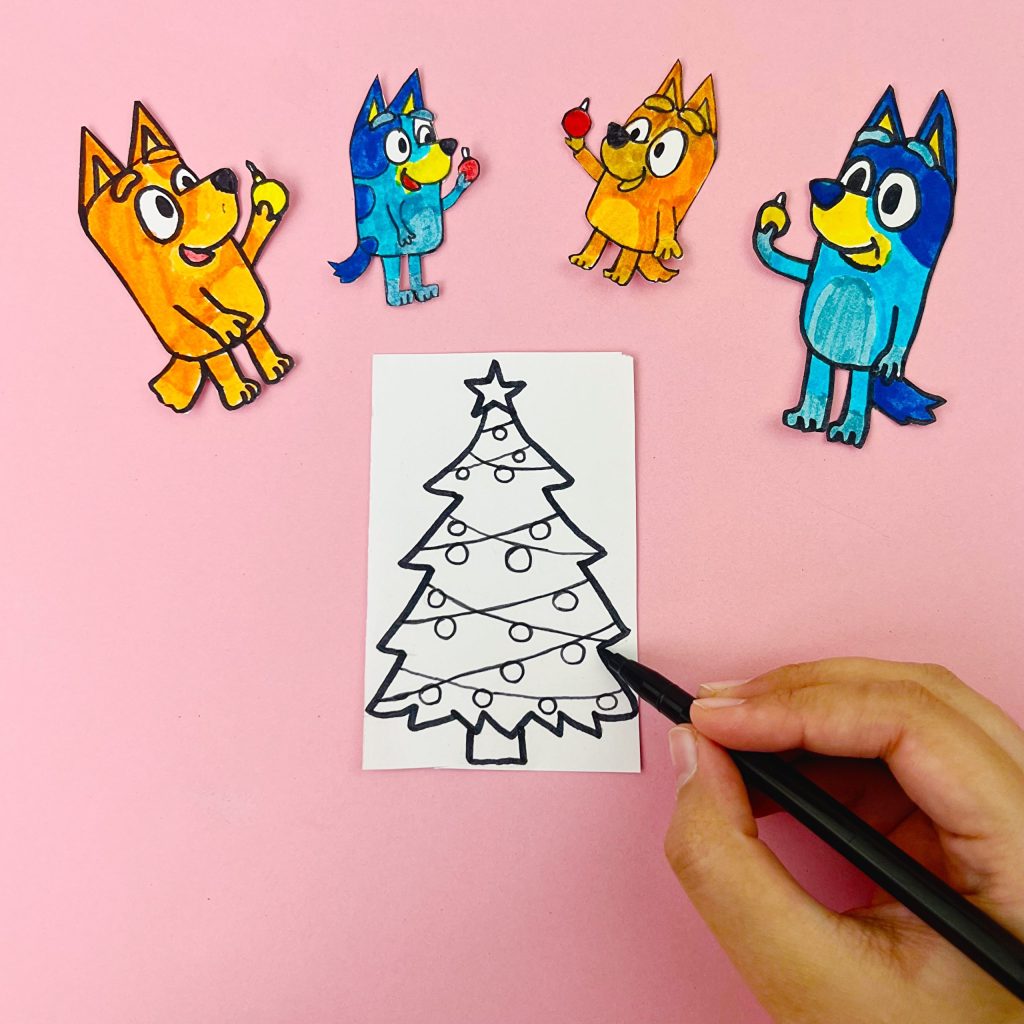 A hand using a black marker to draw a Christmas tree with baubles and a star on white card, surrounded by cut-out Bluey family characters on a pink background, preparing for a holiday craft scene.