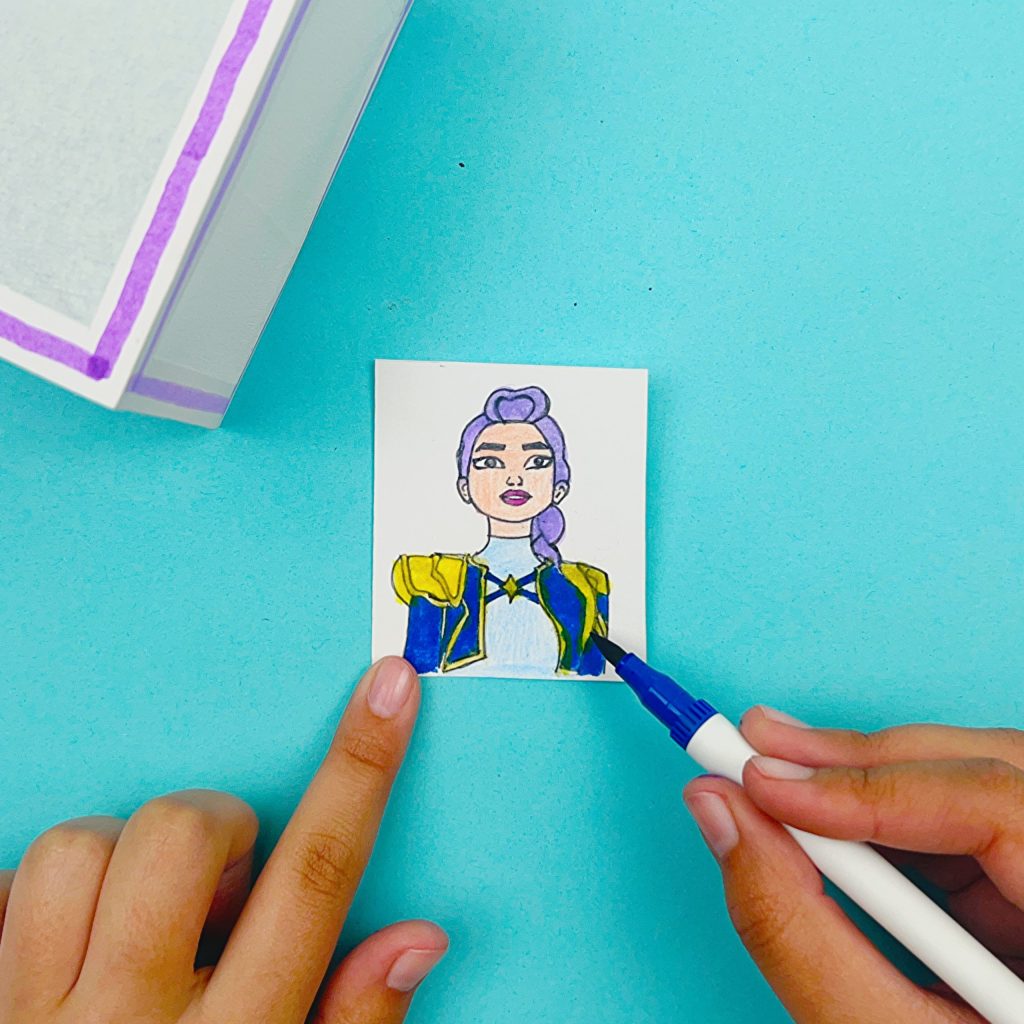 A hand coloring a small cut-out drawing of Rumi from KPop Demon Hunters with markers, filling in her purple hair and blue-and-gold outfit so the character stands out clearly, shown on a blue background beside a paper lantern.